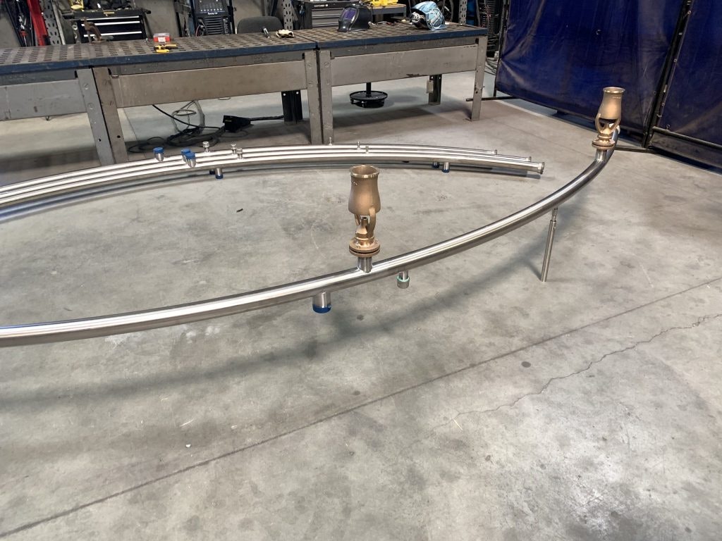 Curved metal pipes with fittings on a workshop floor in front of a workbench.