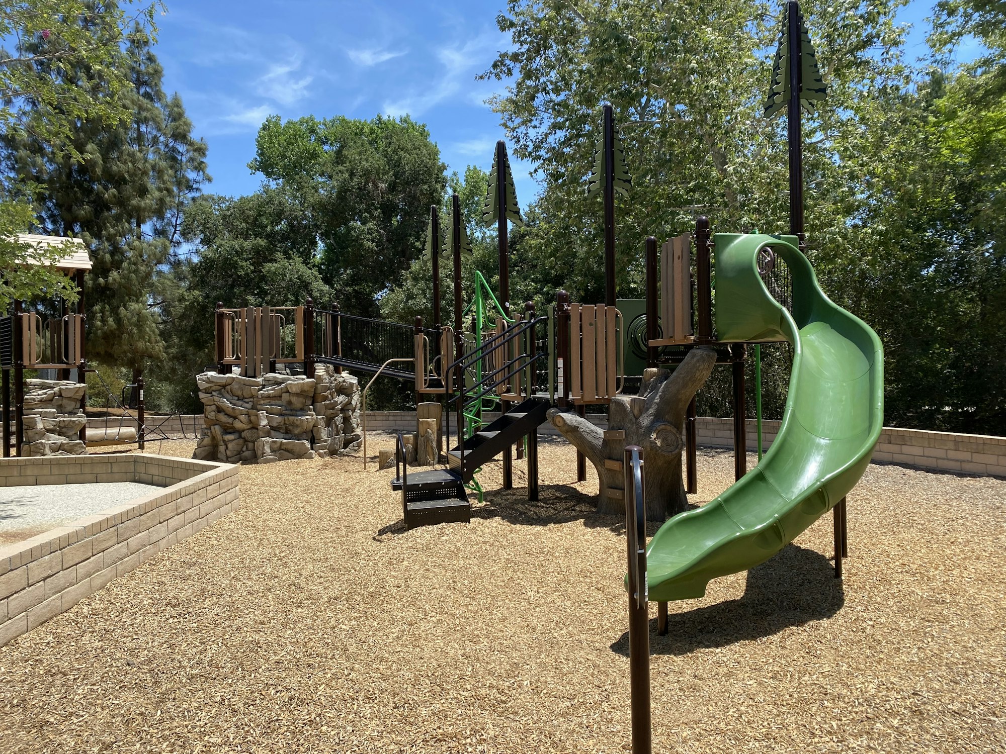 Playground with green slide, climbing structures, and woodchip ground cover, surrounded by trees.