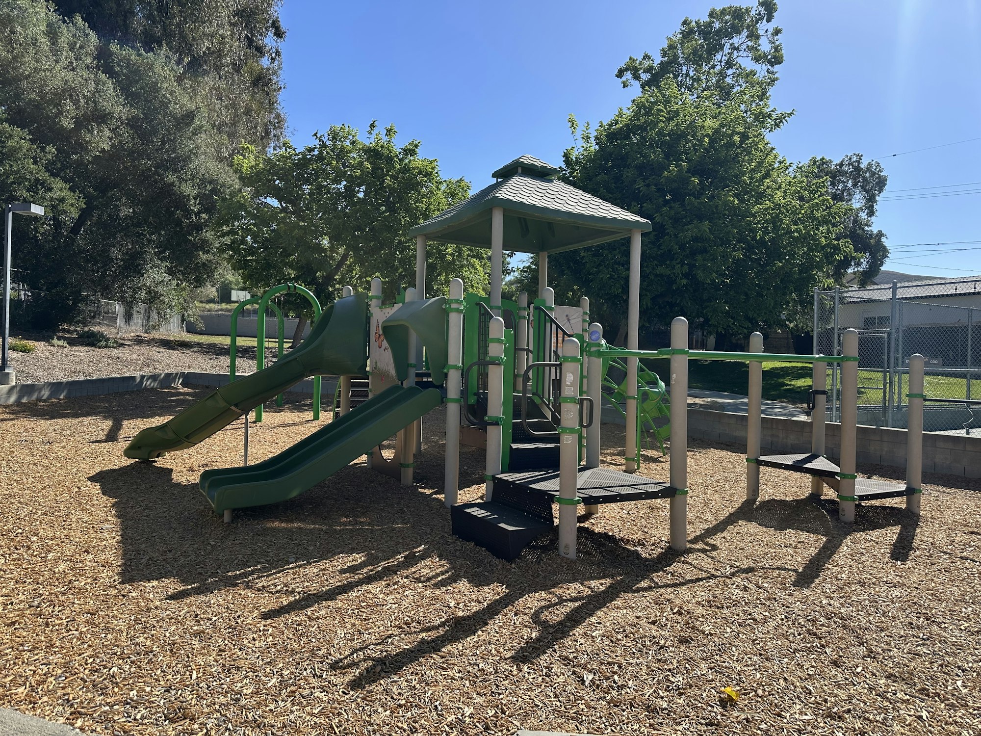 A sunny playground with green slides, climbing structure, and wood chip flooring, surrounded by trees and a fence in the background.