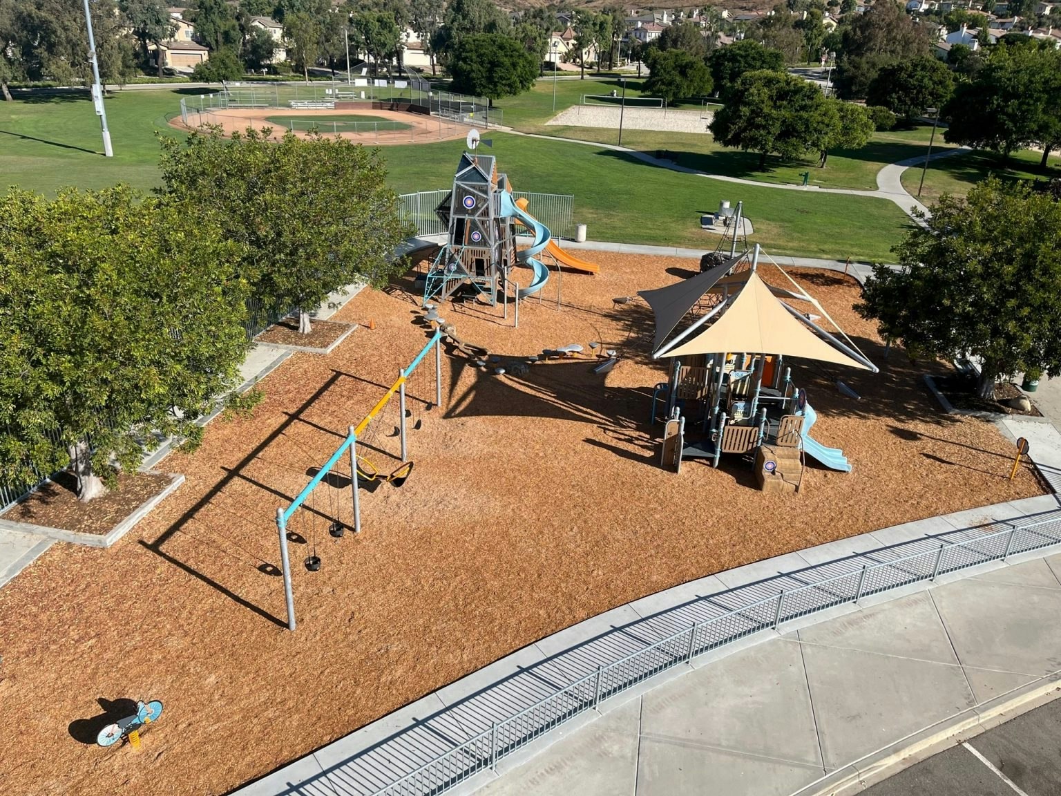 A playground with slides and swings next to a baseball field, surrounded by trees and pathways.