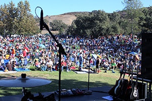 Stage view of a large, seated outdoor crowd with musical instruments and microphone stand visible in the foreground.