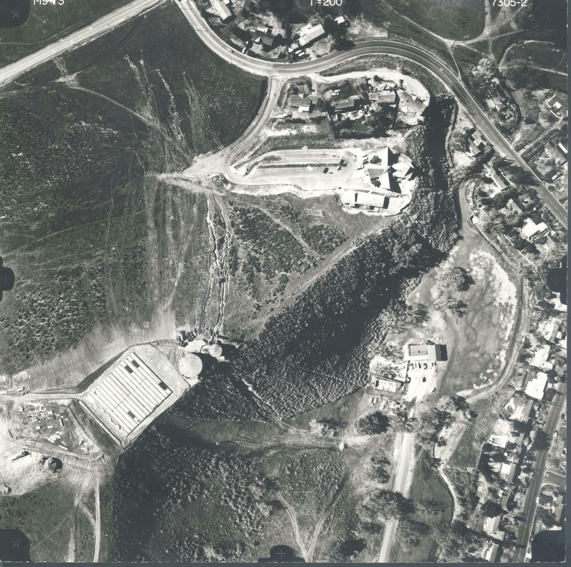 Aerial view of a landscape with roads, buildings, vegetation, and a large rectangular structure.