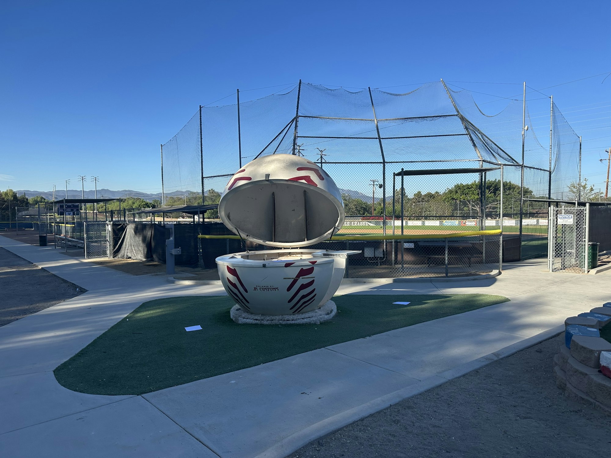 The image shows a baseball batting cage area, featuring a large, open, egg-shaped structure and a clear blue sky.
