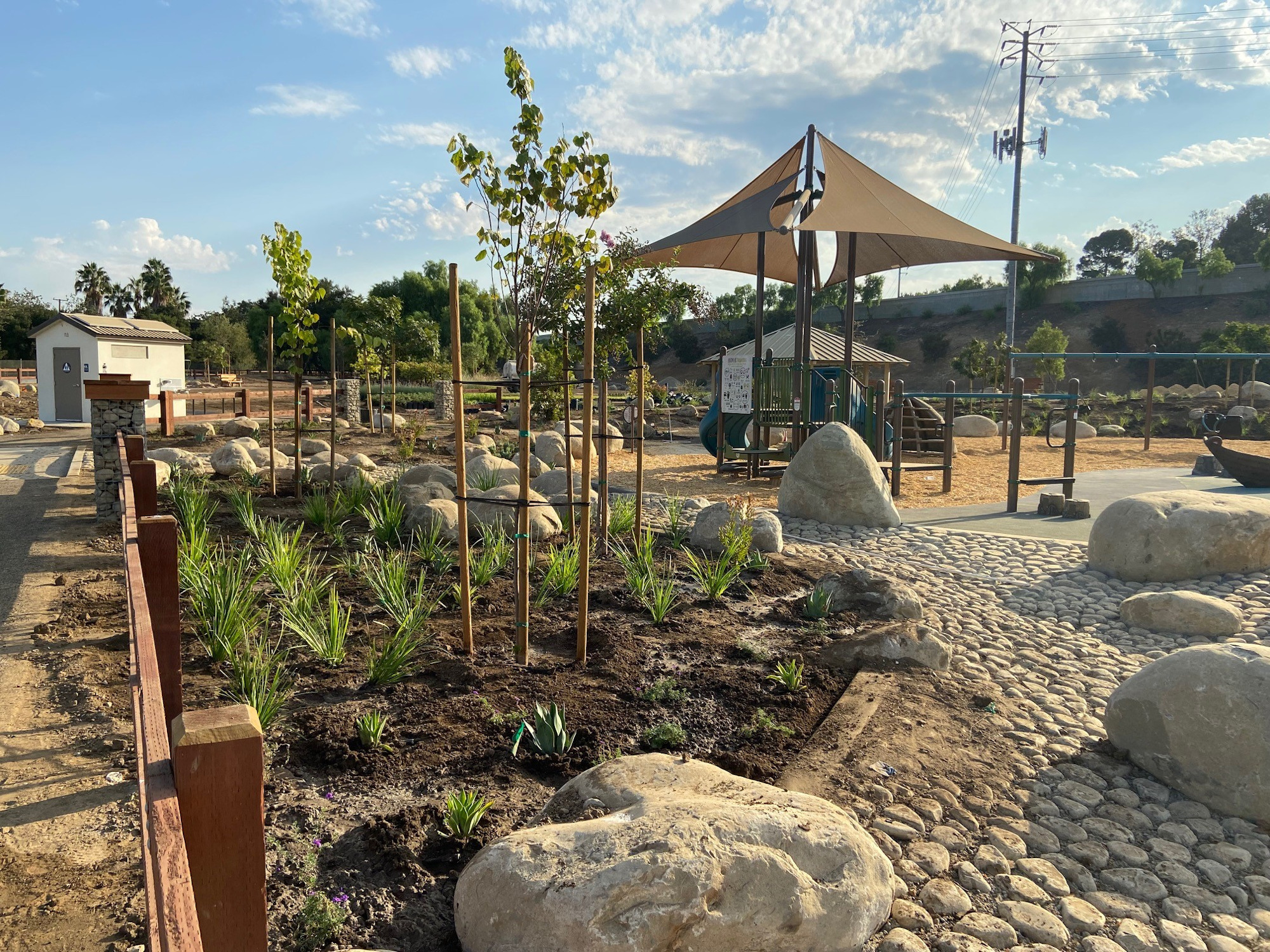 Park with young trees, rocks, a playground, a shaded area, and a restroom building under a clear sky.