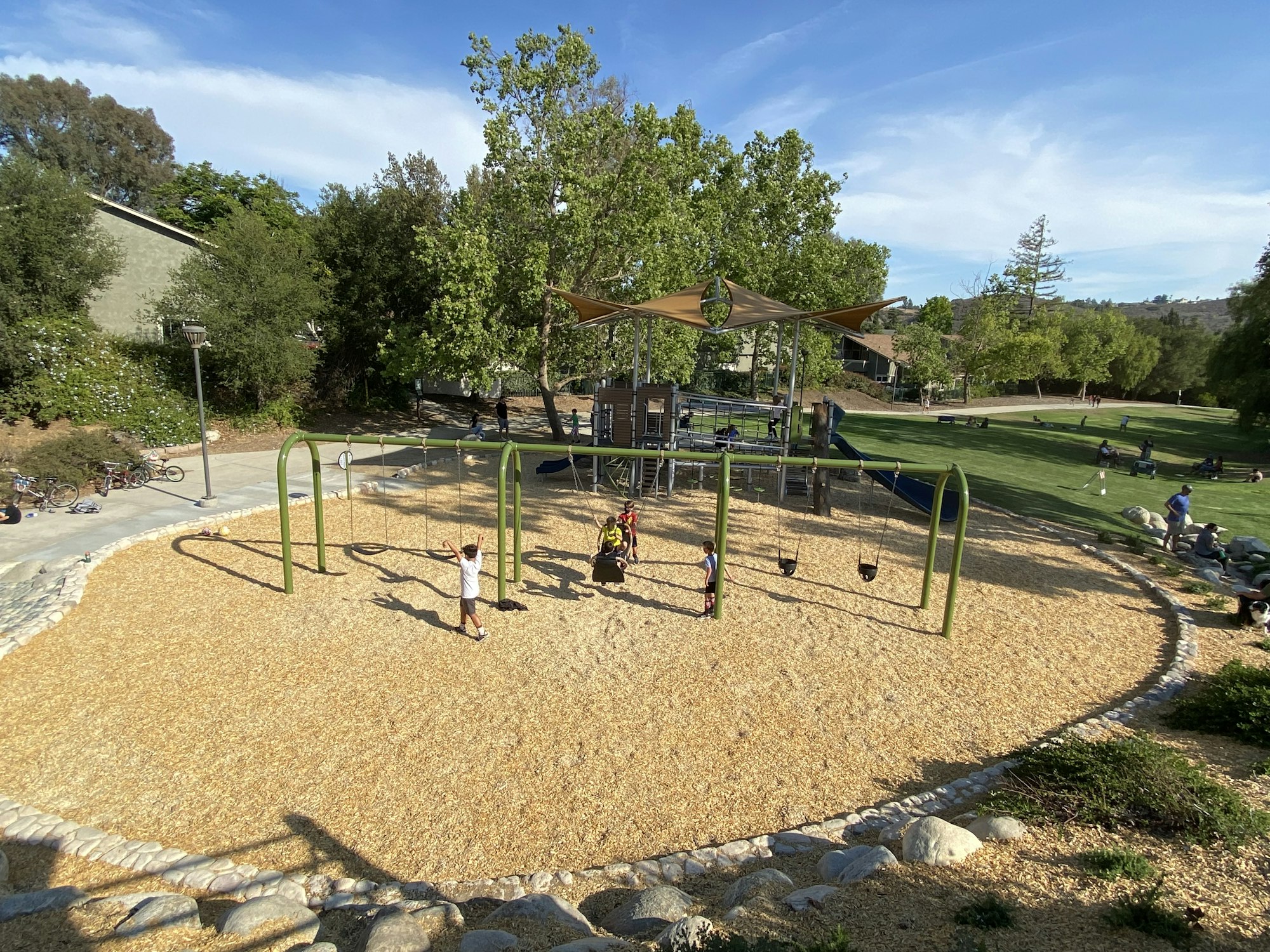 Children playing in a park playground with swings and a climbing structure, surrounded by trees and people on the grass.
