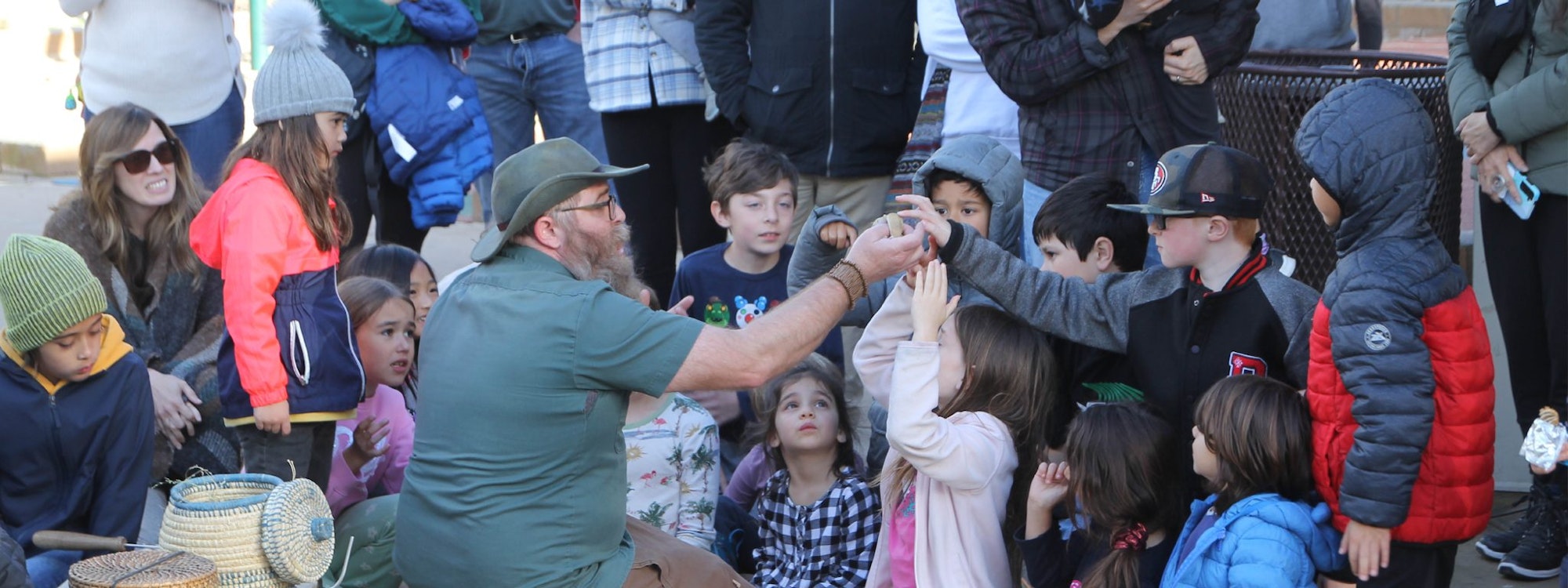 A group of children gathered around a man showing them something, outdoors, with some adults nearby.
