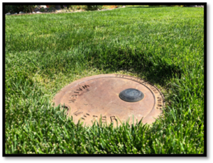 A circular metal access cover resting in vibrant green grass, likely related to water utility access.
