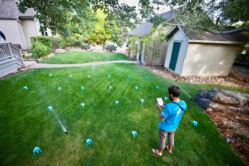 A boy stands in a yard with sprinklers, holding a device, surrounded by green grass and a house in the background.