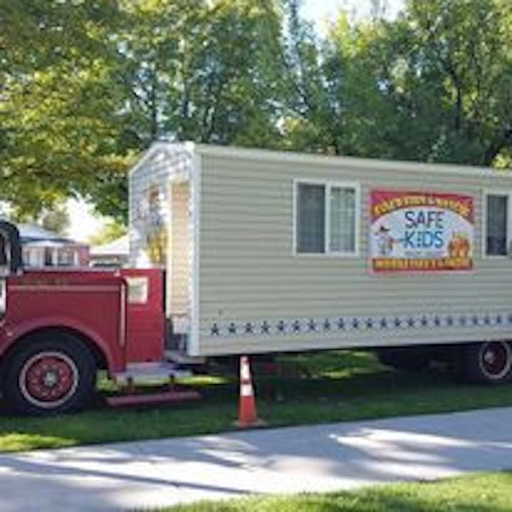 A vintage fire truck pulling a mobile unit with a "Safe Kids" sign, parked on a grassy area with trees around it.