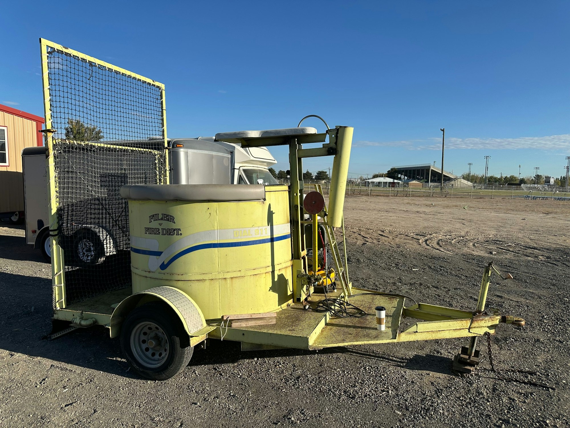 A yellow gel fire suppression trailer with a water tank and equipment is parked on gravel near a building and open field.