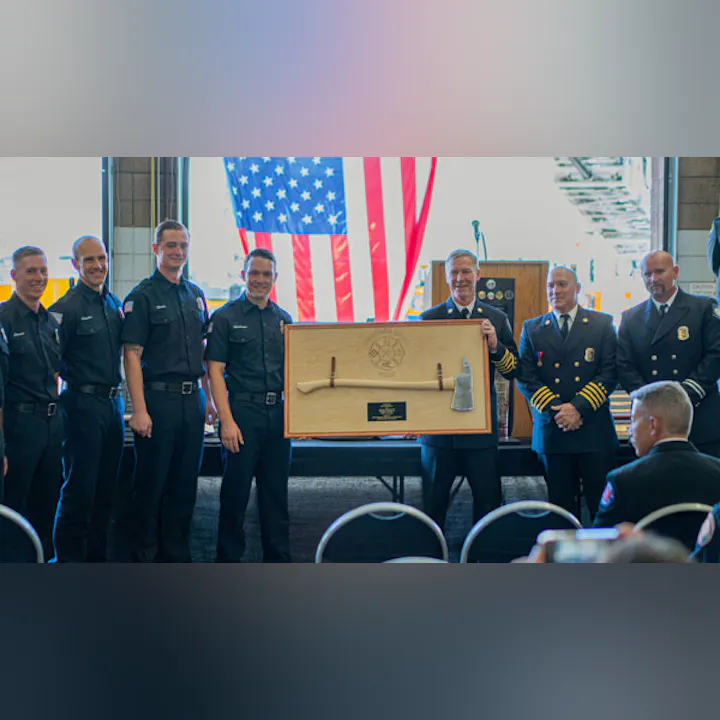 A group of uniformed firefighters holding a ceremonial plaque with an axe, in front of a U.S. flag.