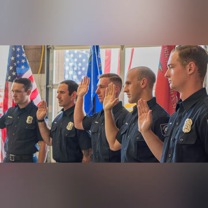 Five uniformed firefighters raising their right hands in a formal setting with flags in the background.