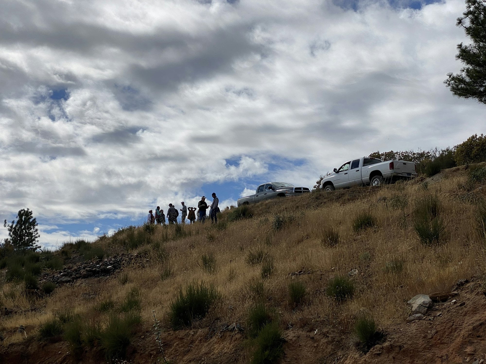 A group of people stand on a grassy hill next to two parked trucks under a cloudy sky.