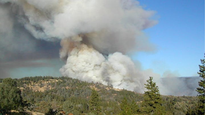 Large plumes of smoke rise over a forested area, possibly from a wildfire.