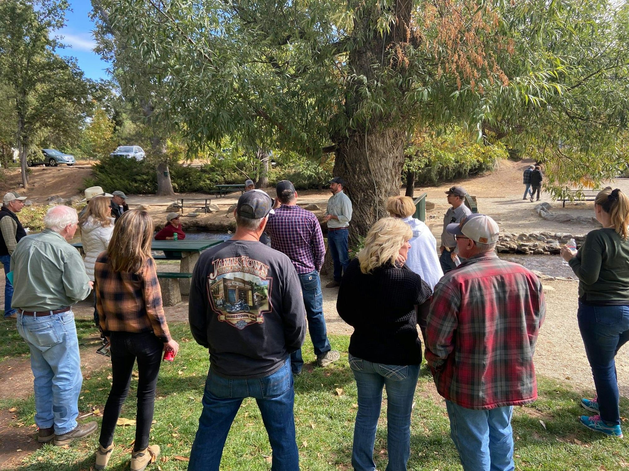 A group of people gathered outdoors near a tree, seemingly engaged in conversation or listening to someone speak.