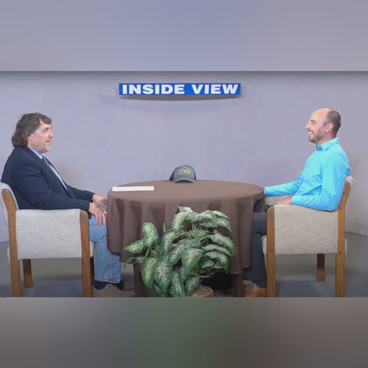 Two men sitting at a round table with a plant in front, "INSIDE VIEW" sign above them, and a UTICA hat on the table.