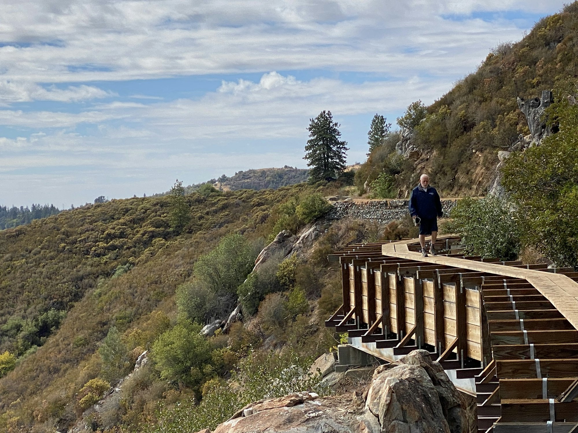 A person walking on a raised wooden path through a hilly, tree-filled landscape under a cloudy sky.