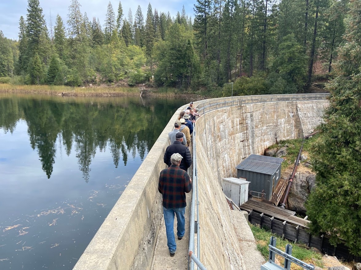 People walking on a curved dam with a lake on the left and trees in the background.