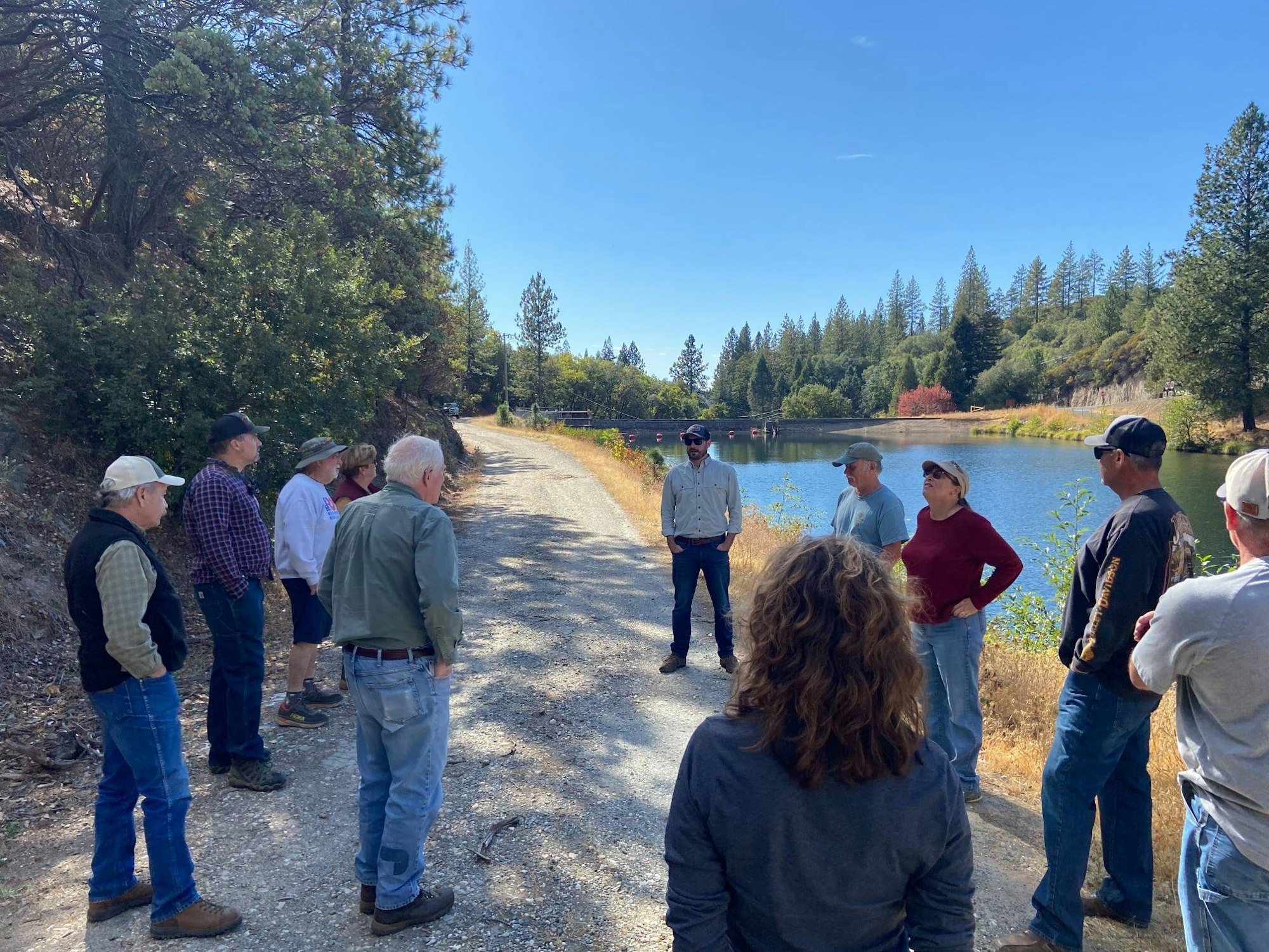 A group of people gathered on a dirt path by a lake, surrounded by trees on a sunny day.