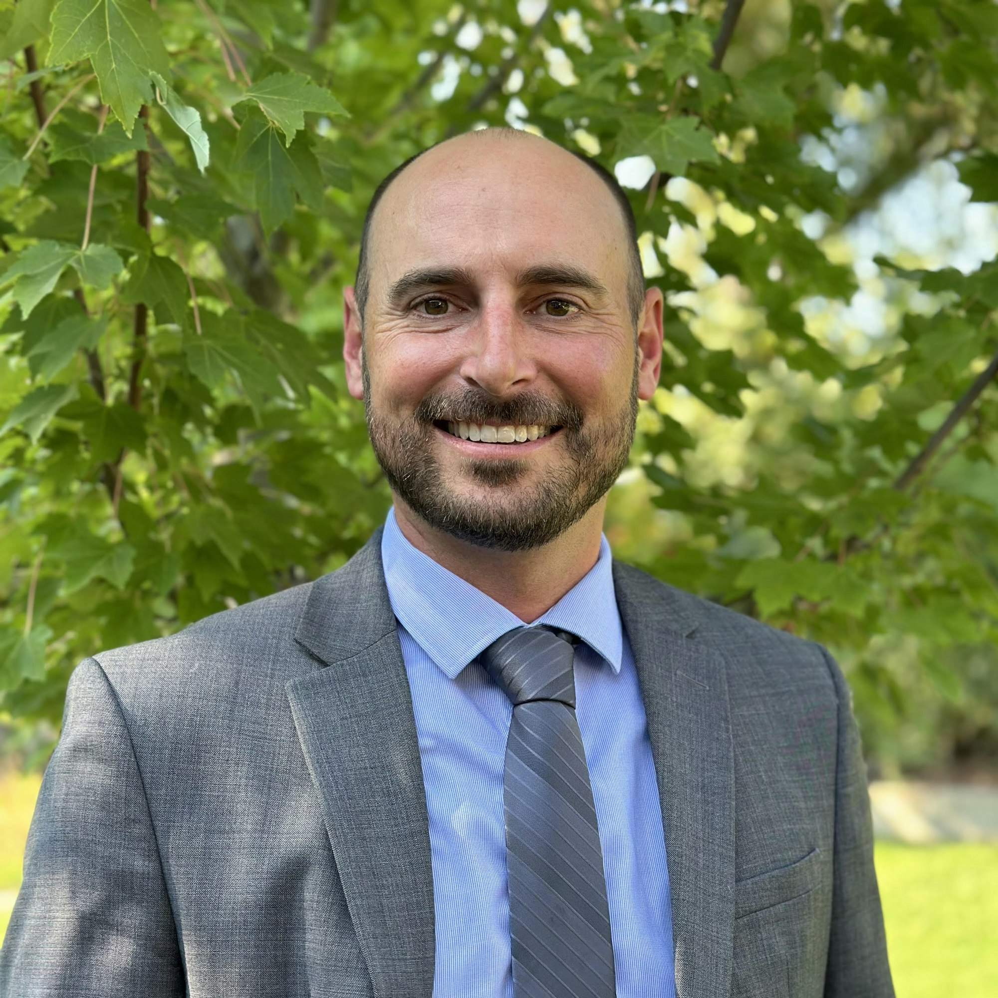 A man in a suit smiling, standing in front of green foliage.