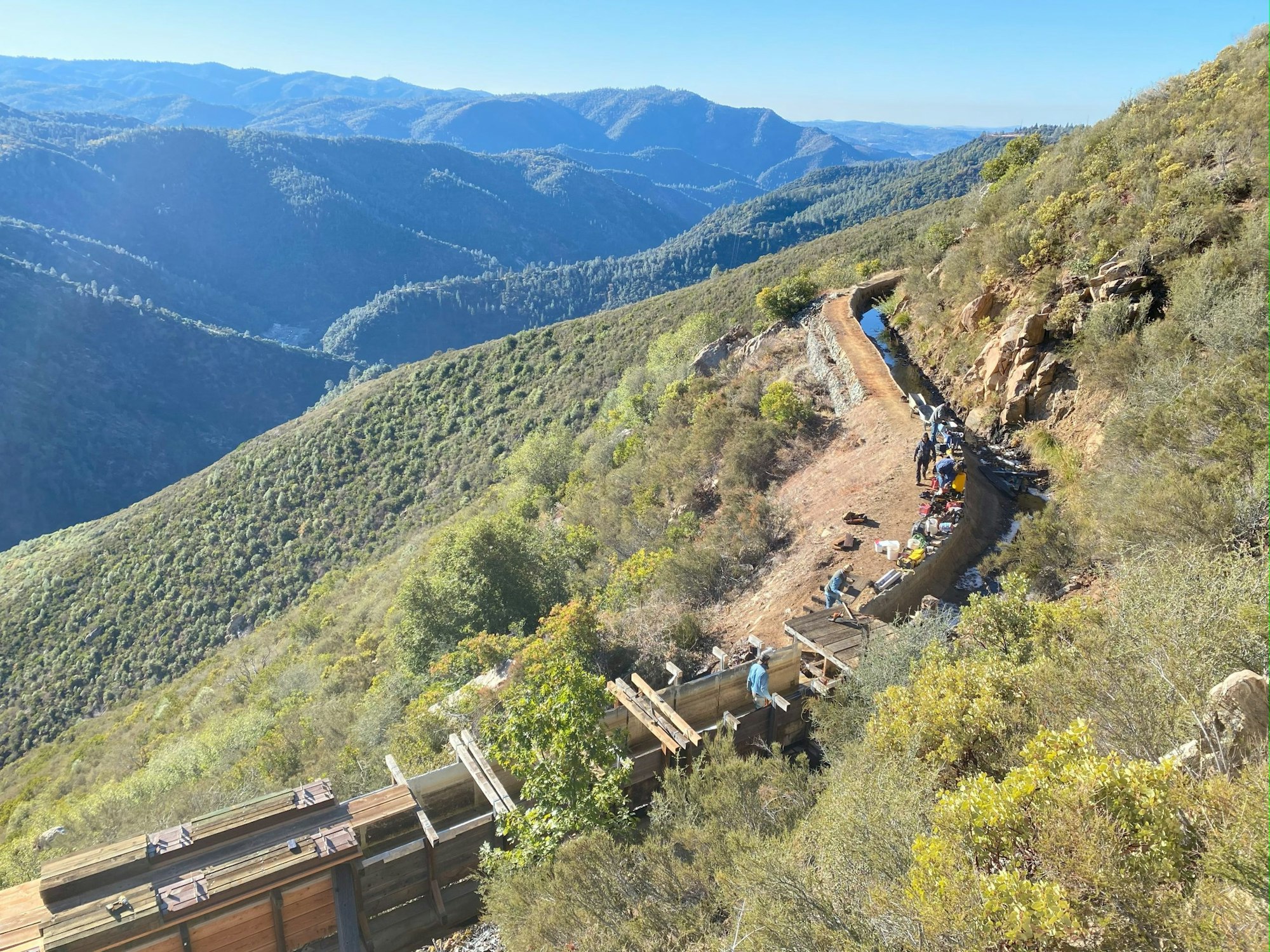 Mountain landscape with a water channel and people working near a wooden structure.
