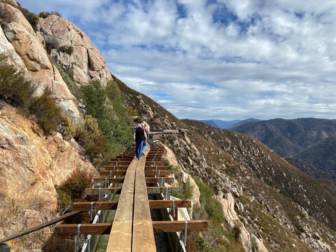 A person walks on a narrow wooden path along a mountainous terrain under a partly cloudy sky.