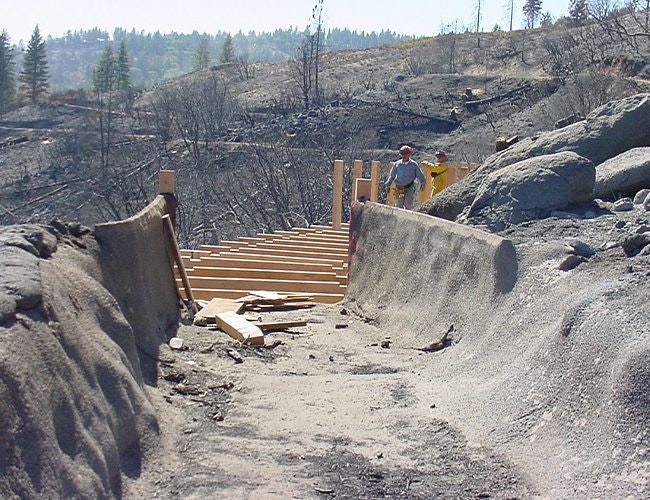 Damaged road with a bridge under construction and workers in a fire-damaged landscape.