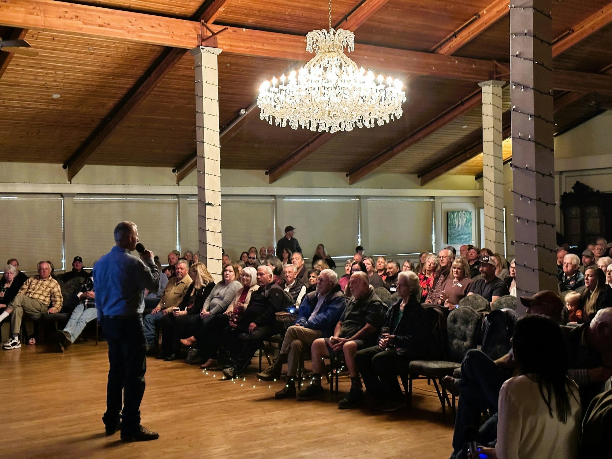 A person speaks to an audience seated in a large room with a chandelier and decorated pillars.