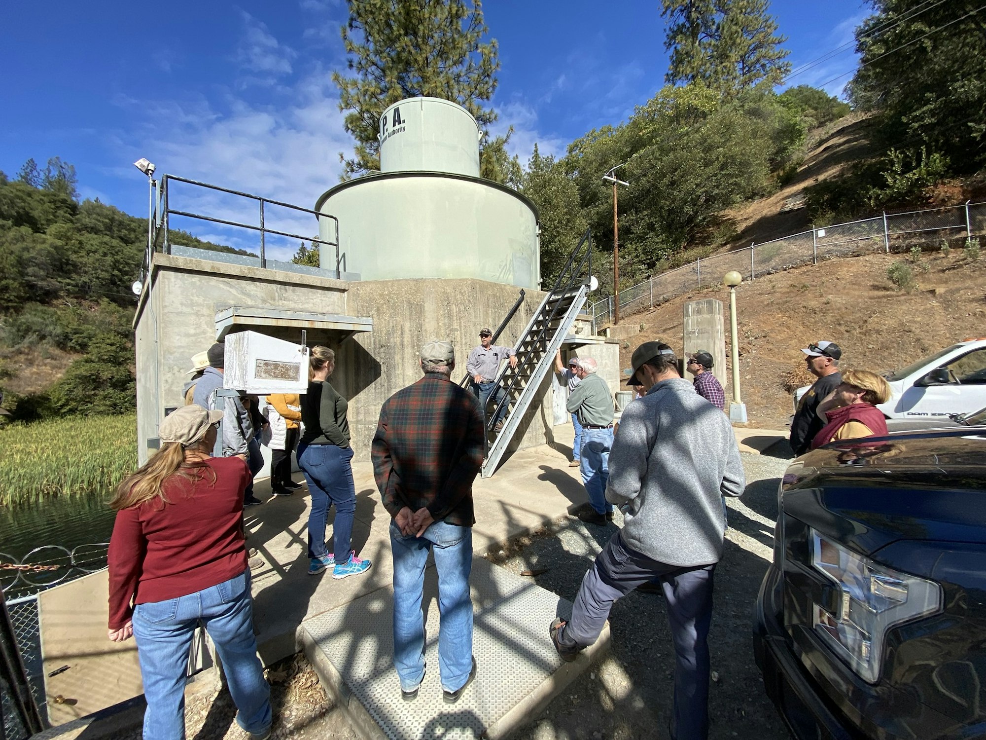 A group of people is gathered outside near a large metal structure, under a clear sky.