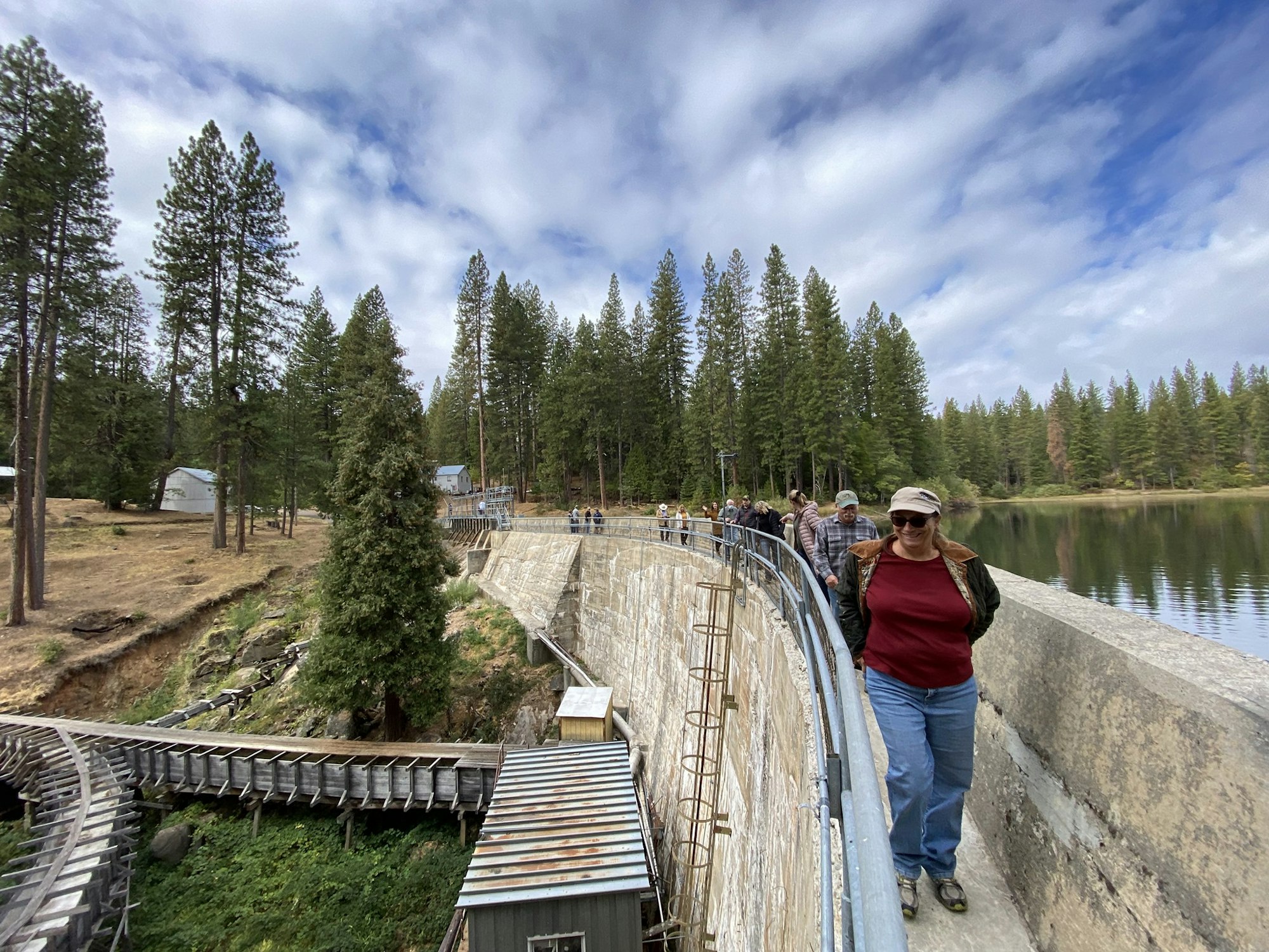 People walking on a concrete pathway beside a forested lake with pine trees under a partly cloudy sky.
