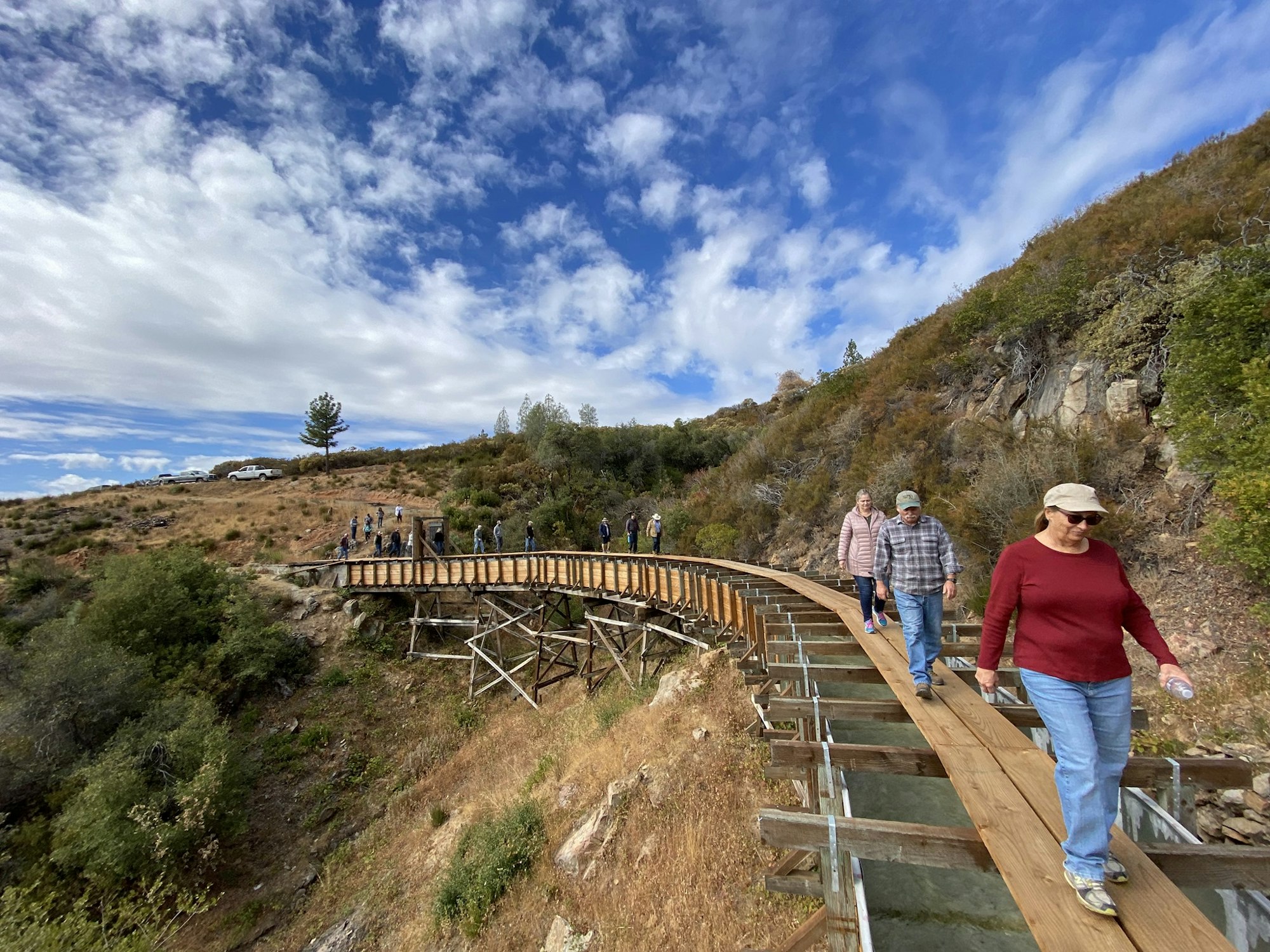 People walking on a wooden bridge with a hilly landscape and cloudy sky.