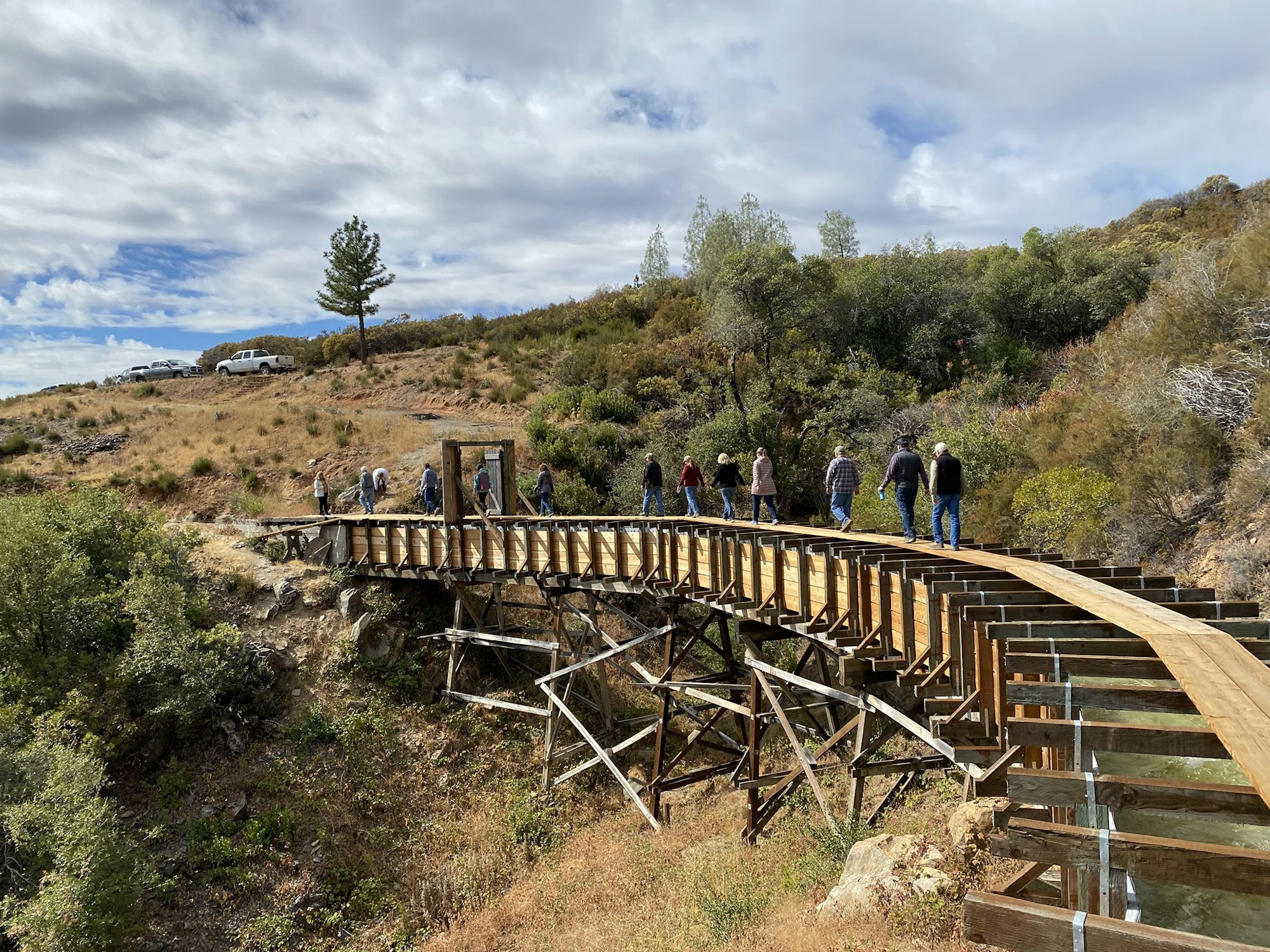 A group of people walking across a wooden bridge in a hilly, rural landscape with cars parked nearby.