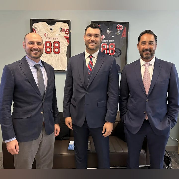 Three men in suits standing in front of two framed Fresno State football jerseys.
