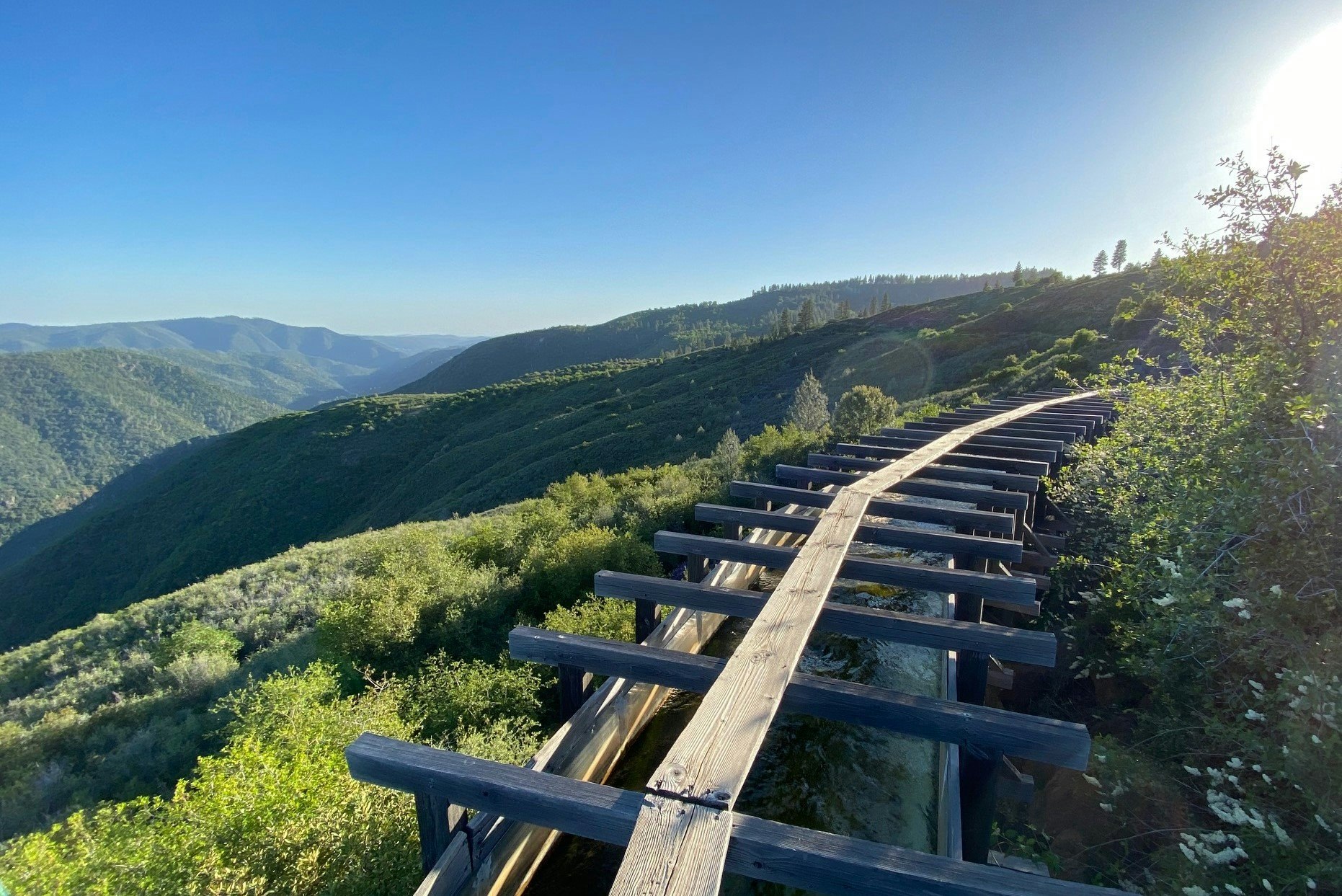 Mountain landscape with a wooden aqueduct path leading into the distance, surrounded by lush greenery under a clear blue sky.