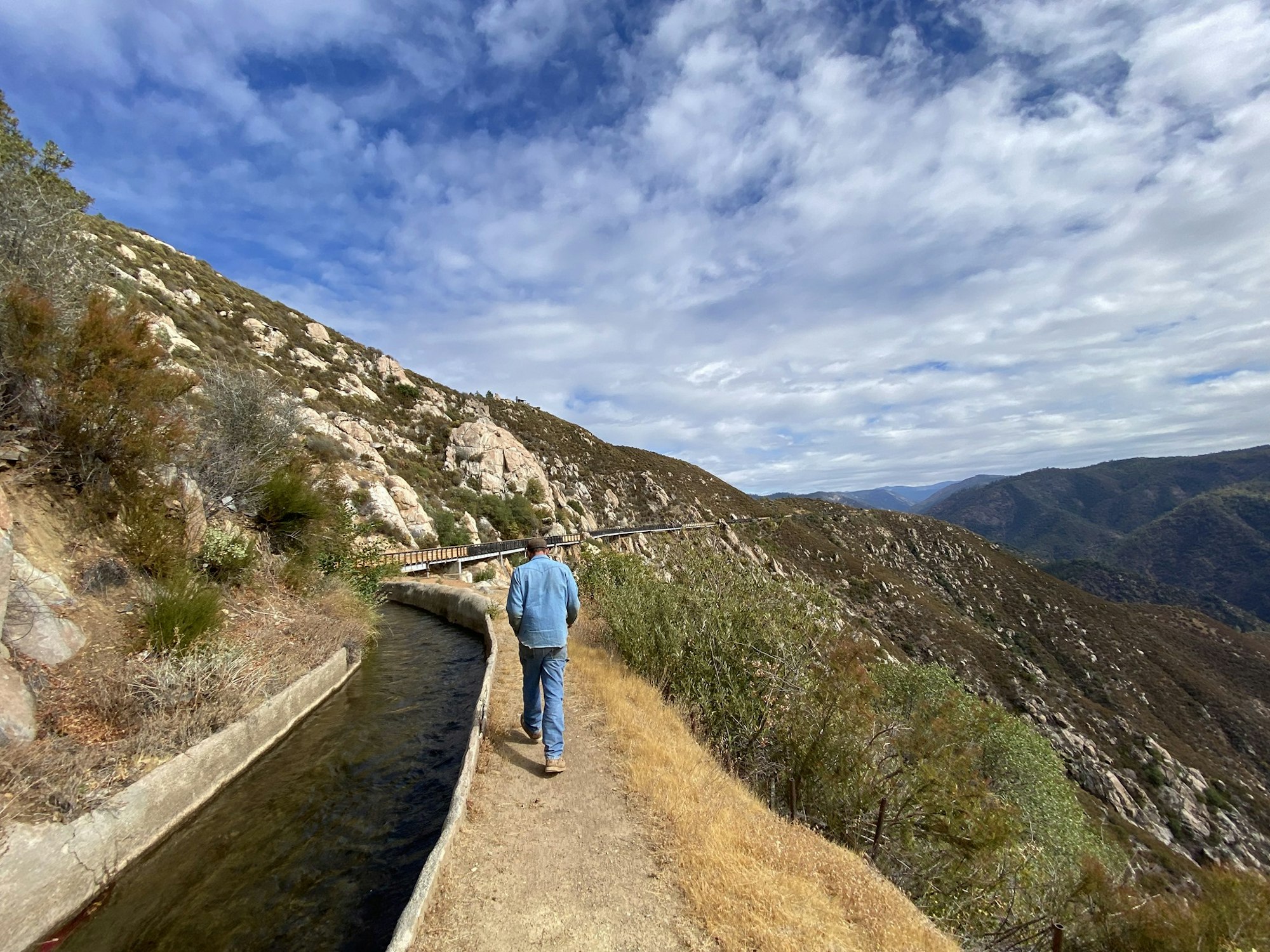 A person walks along a narrow path with a canal on a mountainous landscape under a partly cloudy sky.