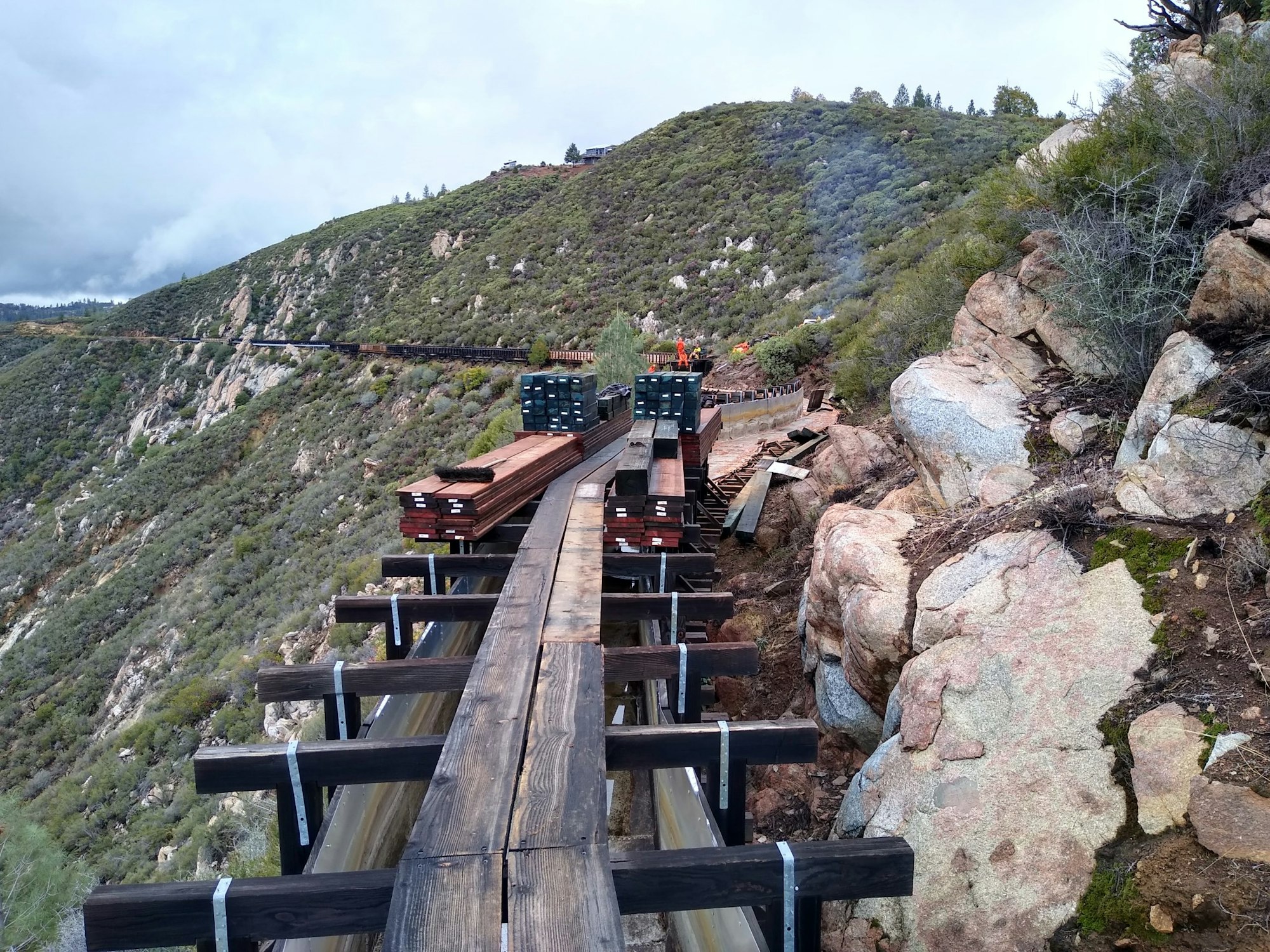 A scenic railway trestle under construction in a mountainous area, with stacks of wooden planks and rocky terrain.