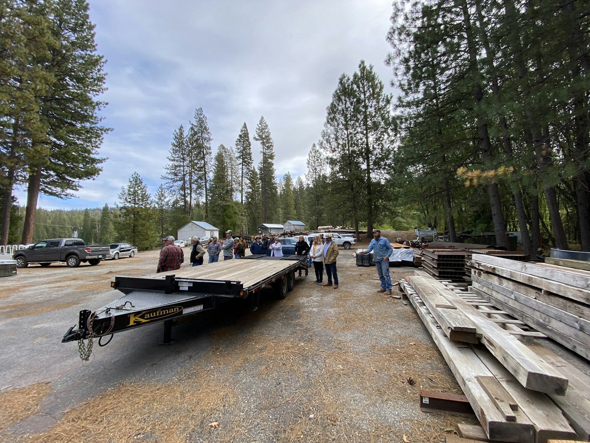 People gathered around a flatbed trailer in a wooded area with trucks and lumber nearby.