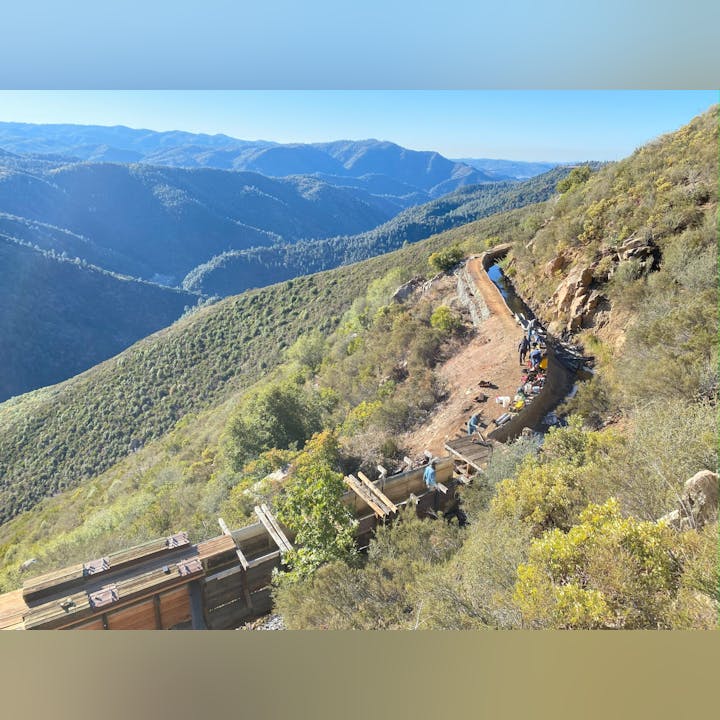 A scenic mountain view with a narrow dirt path along a hillside, where people are working near a ditch or canal.