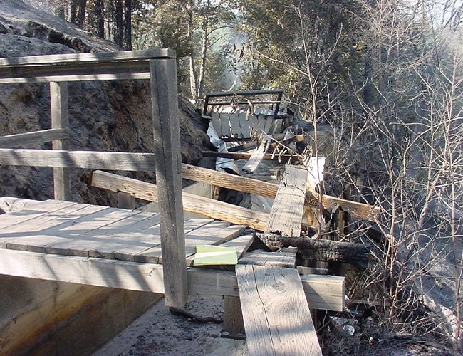 A damaged wooden structure with charred beams in a forest setting, possibly from a fire.