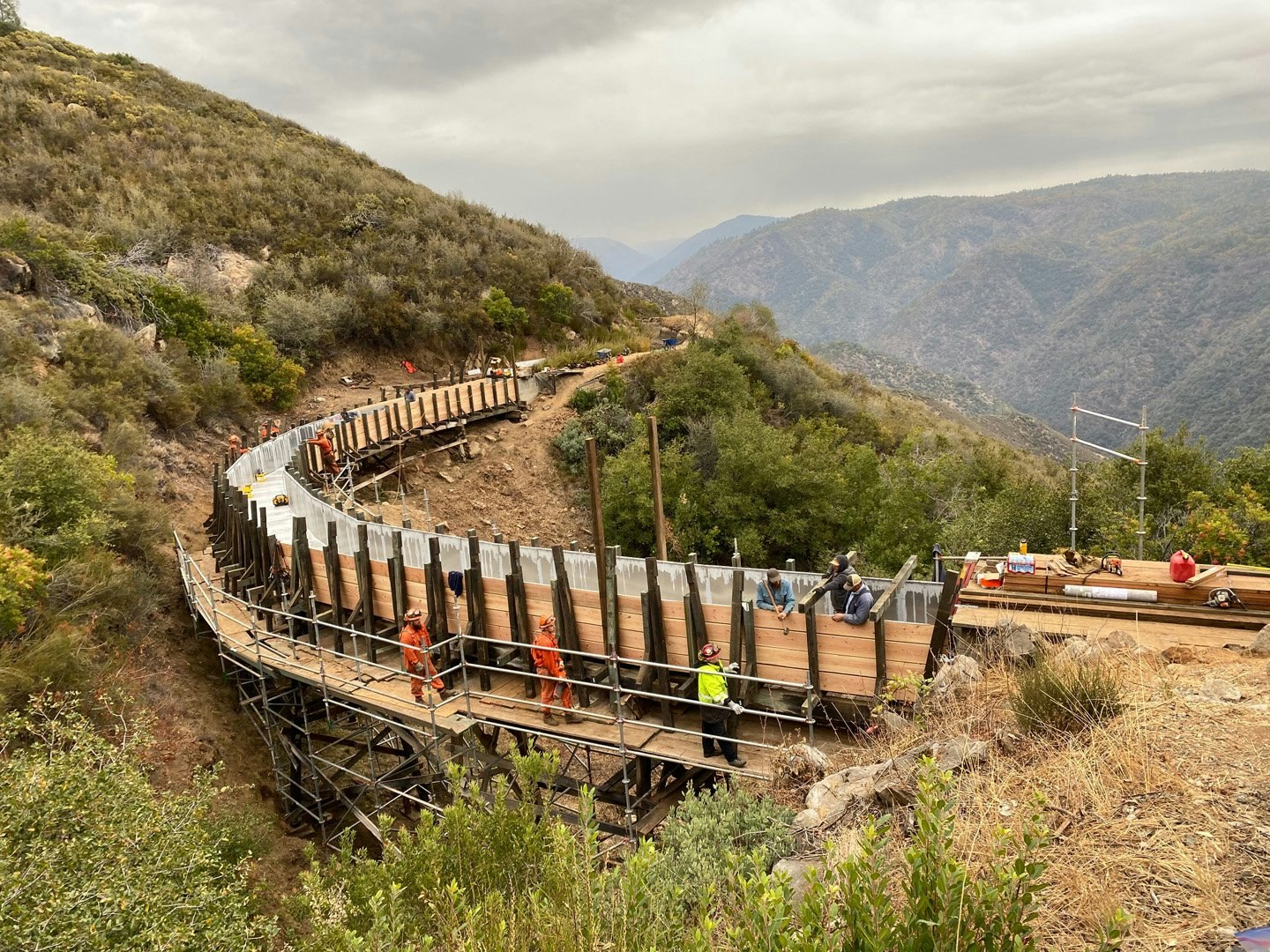Workers on a wooden structure in a mountainous area, constructing a long curved boardwalk with metal supports.