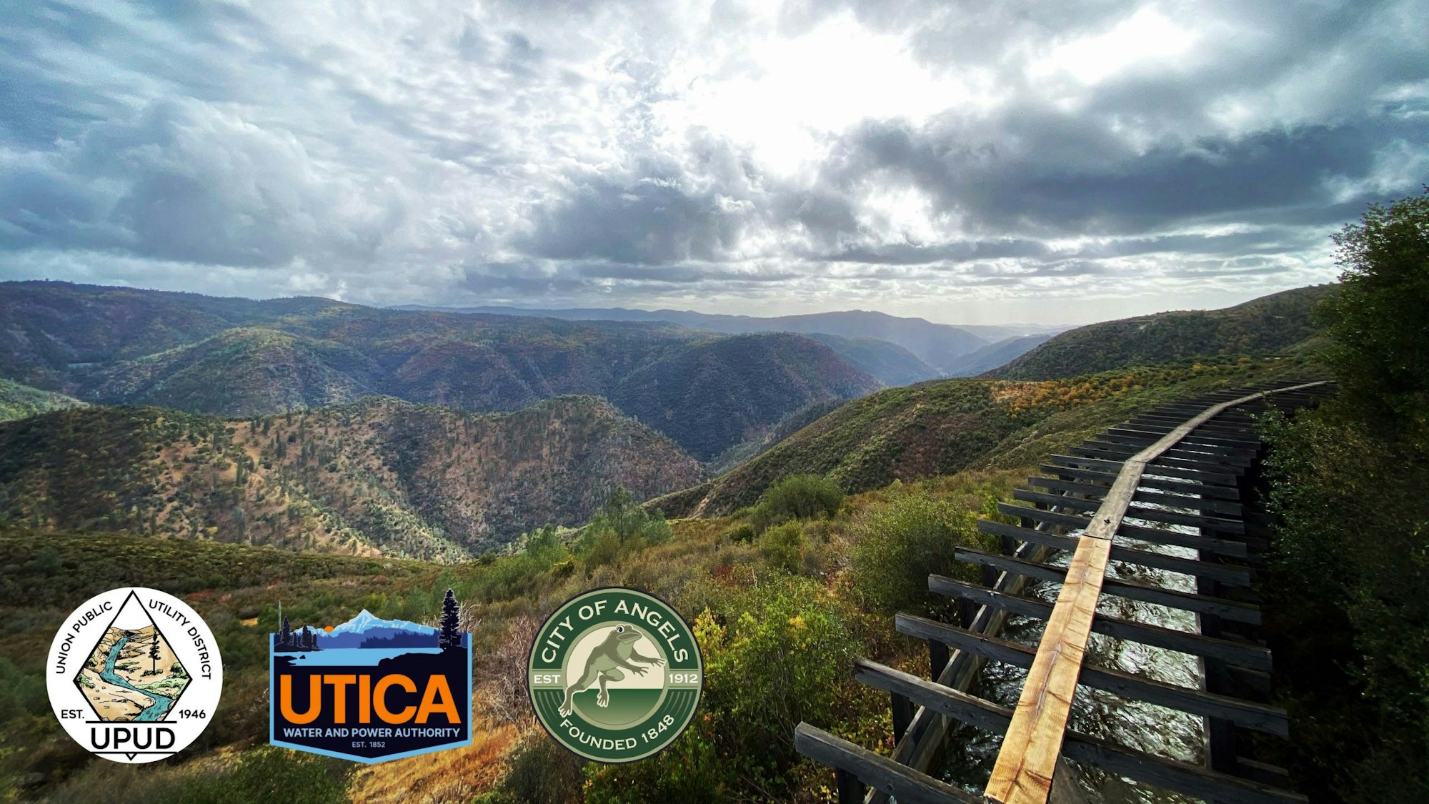 Mountain landscape with cloudy sky, a wooden aqueduct, and logos of UPUD, UTICA, and the City of Angels in the foreground.