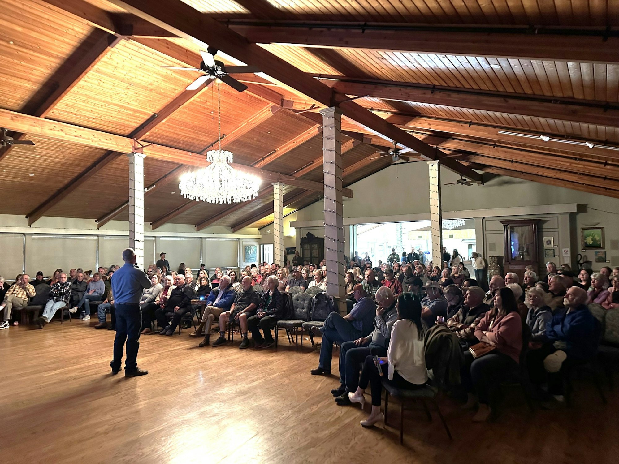 A speaker addresses a seated audience in a large hall with wooden ceilings and a chandelier.