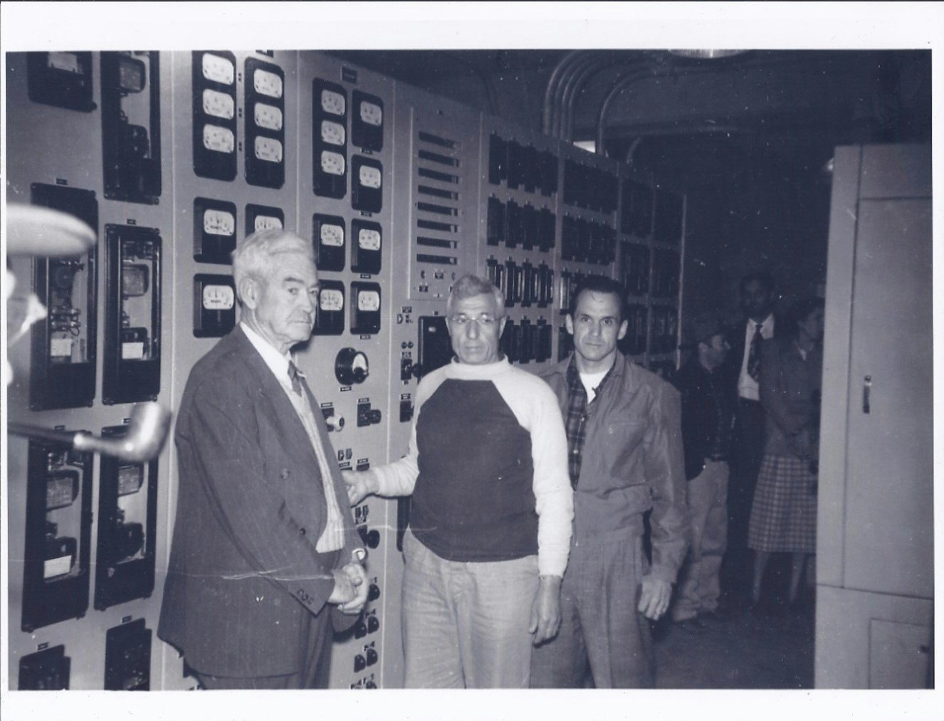 Three men standing in front of a control panel with dials and gauges, in a vintage black-and-white photo.