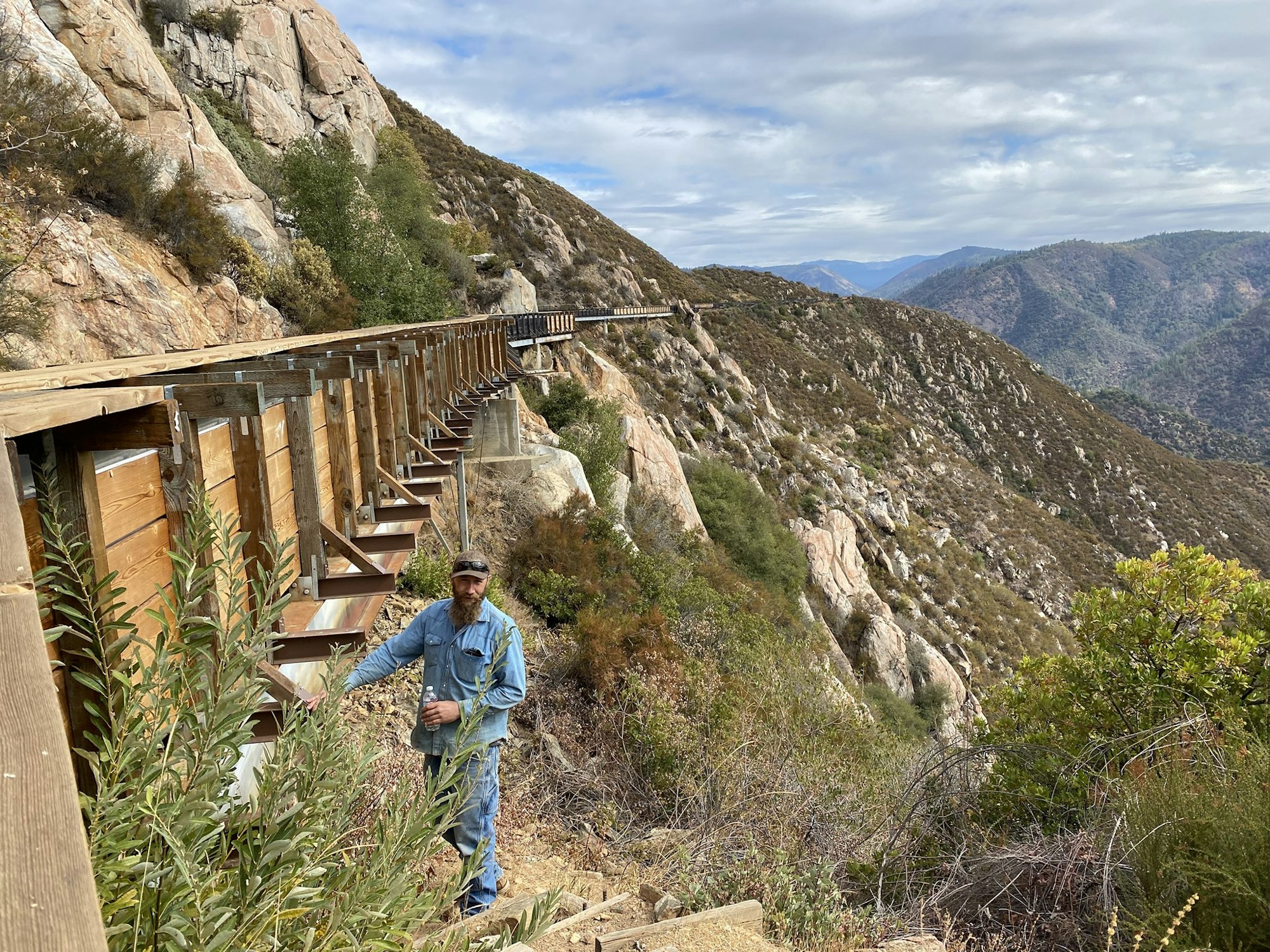 A person in denim clothes walking along a rocky mountainside path with wooden supports, amidst lush greenery and distant hills.