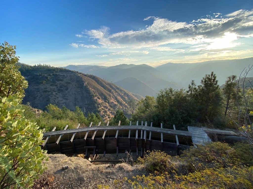 A scenic view of mountains at sunset with trees and a wooden structure in the foreground.