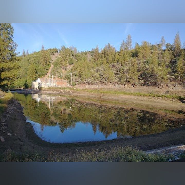 A tranquil pond surrounded by trees and hills, reflecting the clear sky and greenery.