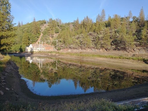 A tranquil pond surrounded by trees and hills, reflecting the clear sky and greenery.