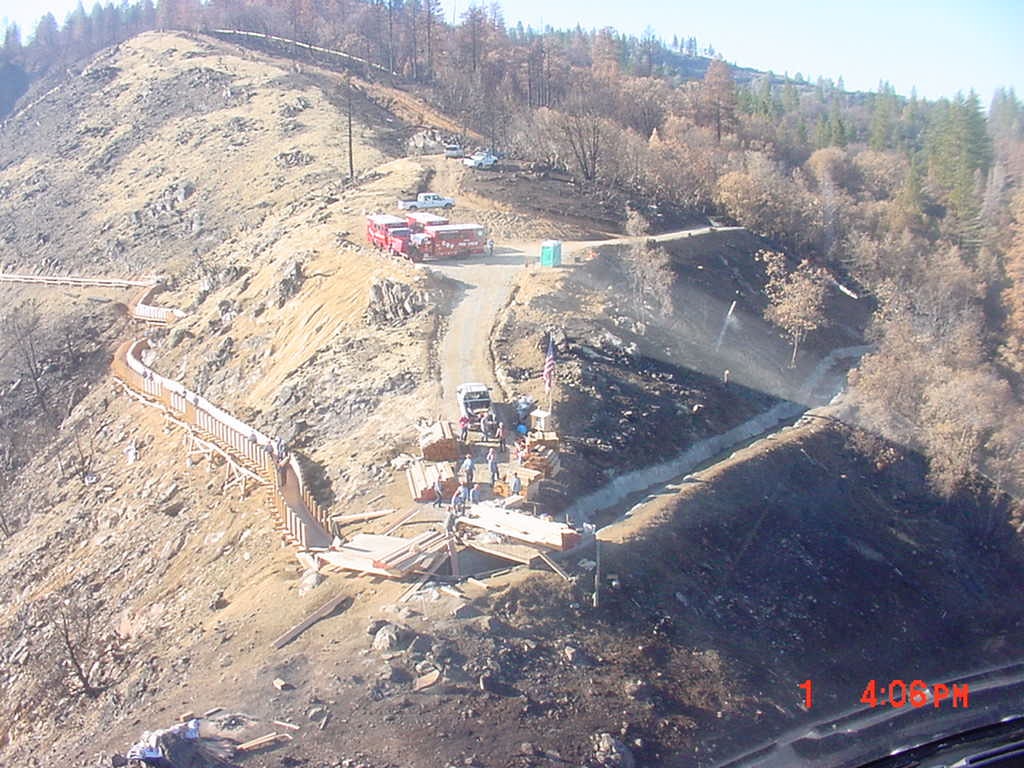 A hillside construction site with vehicles and equipment, surrounded by rocky terrain and forested areas.