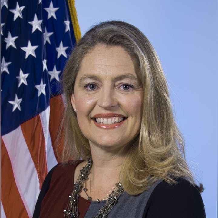 A person smiling in front of the U.S. flag, wearing a necklace and multicolored top, against a blue background.