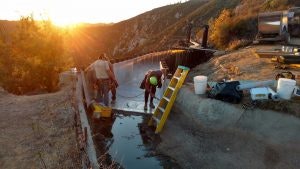 People working near a concrete structure with water at sunset, using a yellow ladder and construction tools around.